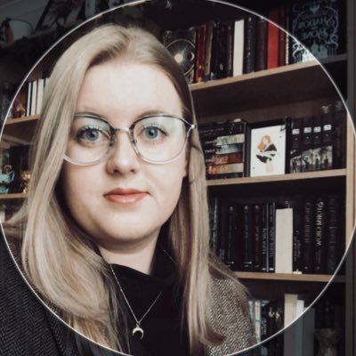 A white woman wearing glasses with long blonde hair, smiling in front of dark bookshelves.